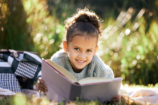 A little girl reading a book while relaxing in a park or garden. Mixed race child learning and getting an education, lying on the grass in nature and having fun