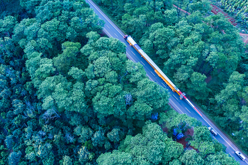 Aerial view of long vehicle truck with special semi-trailer for transporting oversized load or exceptional convoy through a rural forest and mountainous area. Transportation and logistics concept for economics development