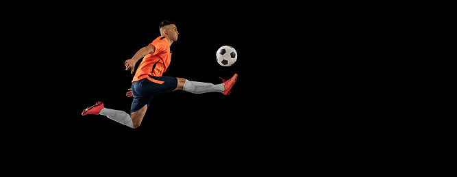 Dynamic portrait of professional male football soccer player in motion isolated on dark background. Concept of sport, goals, competition, hobby, ad. Sportsmen wearing orange-blue football kit