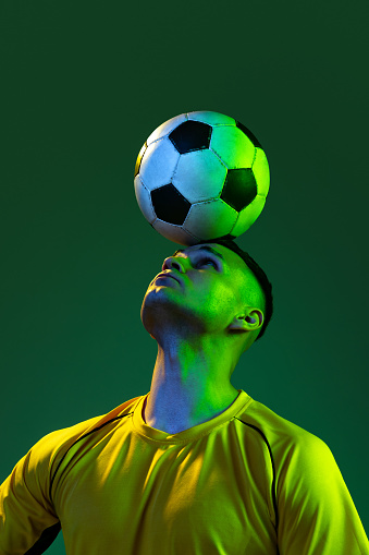 Head pass. Young male football soccer player playing with ball isolated on green background in neon light. Concept of sport, goals, competition, game, achievements. Man in yellow football kit