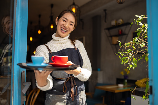 Portrait of cheerful waitress Japanese ethnicity holding tray with coffee and greeting customers