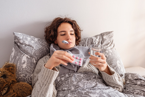 teenage girl has cold, lies in bed covered with blanket, with electronic thermometer, glass of water and blister of antipyretic pills. Viral infection, fever, colds, malaise, flu, runny nose, cough