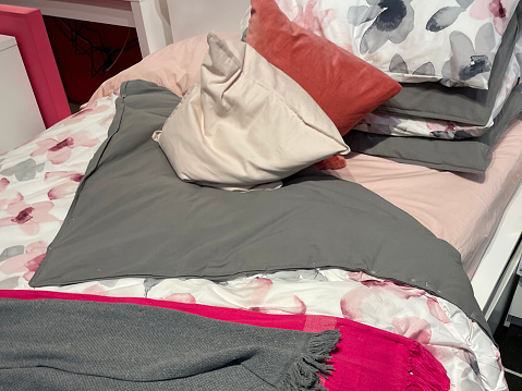 Stock photo showing close-up view of a single bed with a rumpled floral patterned duvet cover, pink and grey blanket throws and scatter cushions.