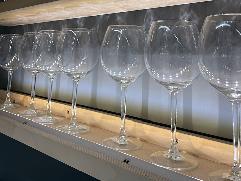 Stock photo showing illuminated, floating shelves displaying rows of long stemmed wine glasses casting shadows on white wall. Home interior design concept.