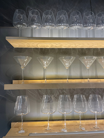 Stock photo showing illuminated, floating shelves displaying rows of martini cocktail and red wine glasses casting shadows on white wall. Home interior design concept.