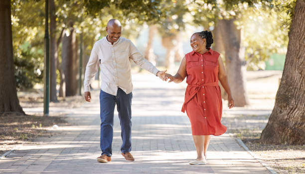 felice coppia afroamericana matura e affettuosa che cammina e si tiene per mano fuori al parco durante l'estate. in amore gli anziani che sorridono mano nella mano mentre trascorrono del tempo di qualità insieme all'aperto - holding hands couple senior couple togetherness foto e immagini stock