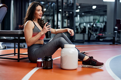 Young fit woman sitting on the gym floor, relaxing and drinking protein shake.