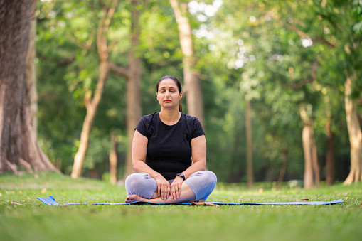 Outdoor image of mid adult woman doing yoga and sitting on yoga mat at park in morning.