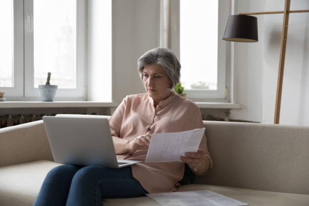 istock Older serious woman sits on sofa with laptop, analyze papers 1400788273
