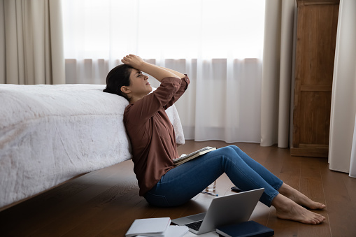 Unhappy stressed young indian ethnicity woman feeling tired of long study time, having problems with understanding educational material, sitting with books and computer on warm floor in modern bedroom