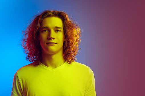 Young attractive man with long curly hair posing isolated on gradient blue-pink background. Concept of beauty, fashion, youth culture and emotions. Half-length male portrait. Copy space for ad