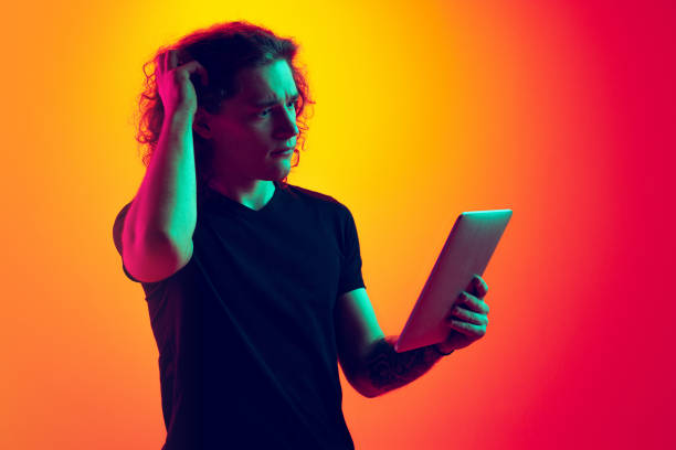 istock Emotive man, student with long curly hair using tablet isolated on gradient yellow-magenta background in neon. Concept of education, fashion, youth culture and emotions 1400791894