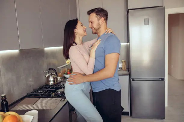 Young Caucasian just-married couple in kitchen of new home. Wife and husband hugging. In love.