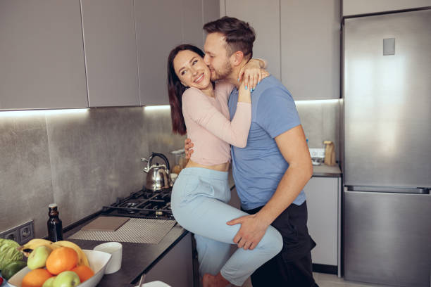 Caucasian handsome man kissing beauiful woman while couple standing in kitchen. Caucasian handsome man kissing beauiful woman while couple standing in kitchen in the morning. trophy wife stock pictures, royalty-free photos & images