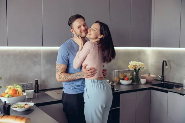 Cheerful just-married young Caucasian couple in hugs having fun and laughing in kitchen. In love.