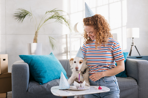 Young happy redhead woman celebrating a birthday at home with her pet dog, holding it in her arms and sitting on the floor