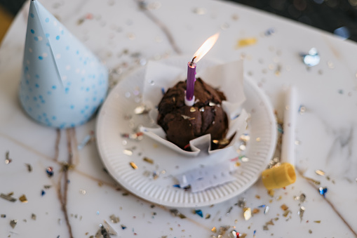 Close up of a small and modest cupcake acting as a birthday cake on a table with a birthday candle in it, a party hat and a party whistle next to it, confetti, no people