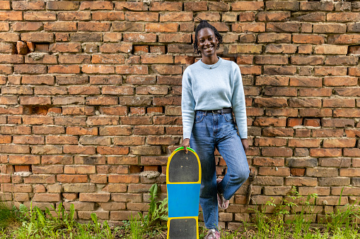 Portrait of young urban woman Black ethnicity in front of the brick wall, holding a longboard