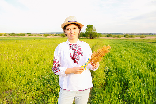 Defocus smile woman in vyshyvanka holding bouquet of ripe golden spikelets of wheat tied on meadow nature background. Flag Ukraine. Independence. Agriculture Ukraine. Support. Freedom. Out of focus.