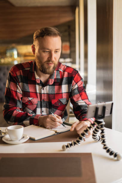 istock Handsome bearded businessman sitting at wooden table with cellphone and writing down notes in notepad, smartphone on tripod online webinar, video blog recording, blogger 1400799616