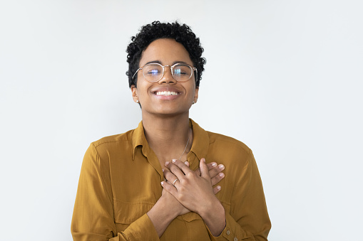 Happy grateful young African girl posing isolated on white, applying hands to chest, thanking, feeling gratitude. Young Black woman in glasses making sign of kindness, showing love, care, support