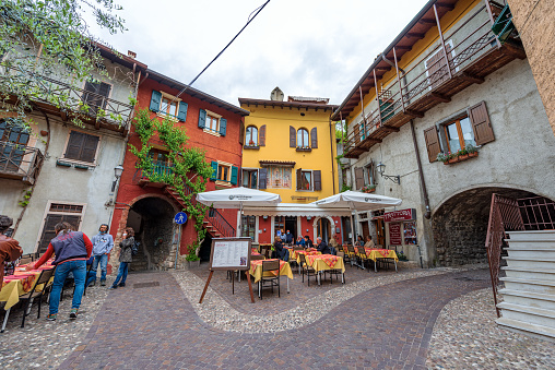 Castelletto di Brenzone, Italy - May 16, 2020: Small restaurant in the small and ancient village of Castelletto di Brenzone, tourist resort on the coast of Lake Garda, Brenzone sul Garda municipality, Verona province, Veneto, Italy, Europe. A group of people have lunch in the small village on a spring day with an overcast sky.
