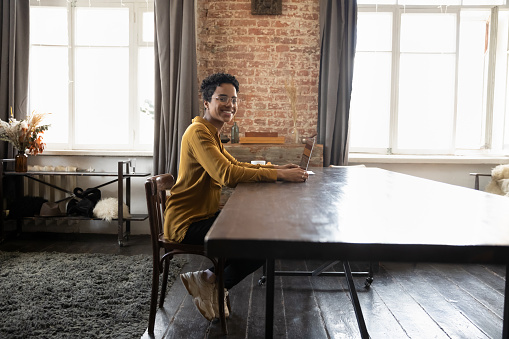 Happy young Black business woman, freelance employee, entrepreneur working at laptop in home office, sitting at big table, looking at camera, smiling. Full length portrait, side view
