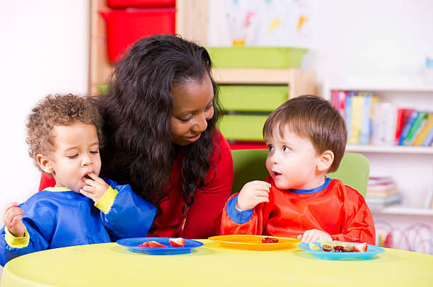 Children eating fruit at a nursery with their carer A stock photo of toddlers eating fruit and having a chat in the playroom. daycare preschool Lunchbox Challenges babies stock pictures, royalty-free photos & images
