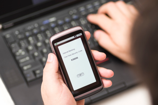 A stock photo of a one time security code on a smartphone. A man receives a one time Security code delivered by SMS and enters it on to a laptop.