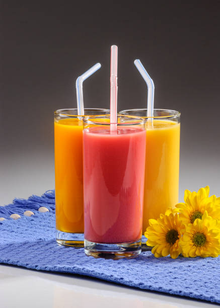 Three glasses of fruit juice with plastic straws over blue placemat on gray gradient background.