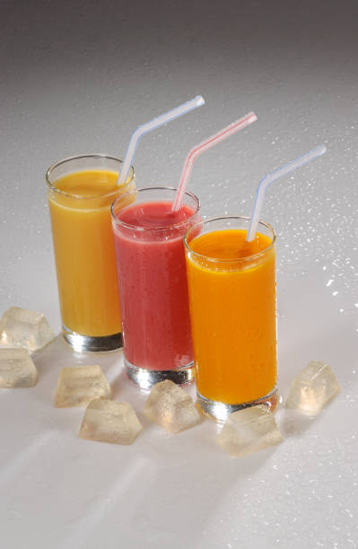 Three glasses of fruit juice with plastic straws and ice on gray gradient background.