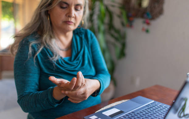 Overworked mature woman with gray hair and wrist pain working on laptop from home, carpal tunnel syndrome stock photo