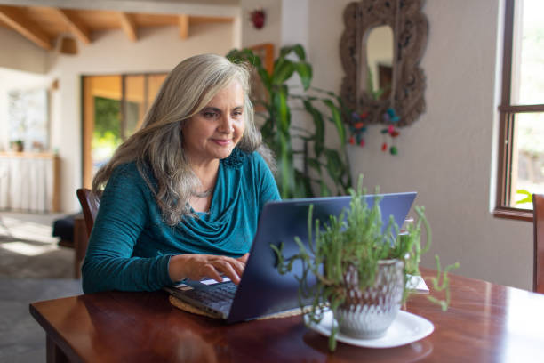 Mature woman with long gray hair  working on laptop from home, smiling stock photo