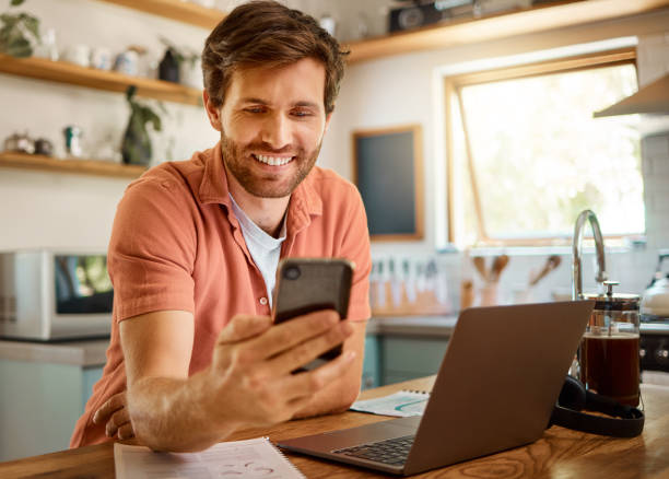 istock Young cheerful caucasian businessman using a phone while working on a laptop at home alone. Happy male businessperson smiling and using social media on a cellphone while working in the kitchen at home 1400808847