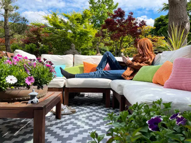 Photo of Close-up image of attractive, red haired young woman lounging on outdoor sofa area in Summer, hardwood seating with cushions, wooden table top with flowering plant centrepiece, Japanese maples, landscaped oriental design garden, sunny day