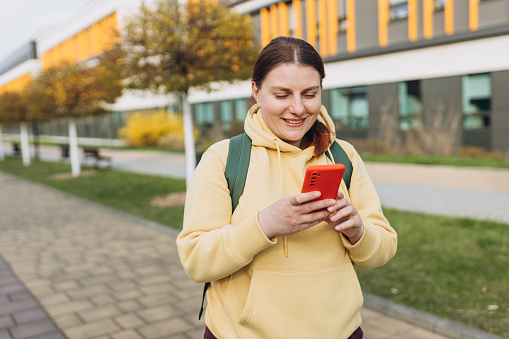 A portrait of a smiling beautiful woman texting sms with her phone on urban background. Happy student with backpack is using a smartphone outdoors, spring or autumn time.