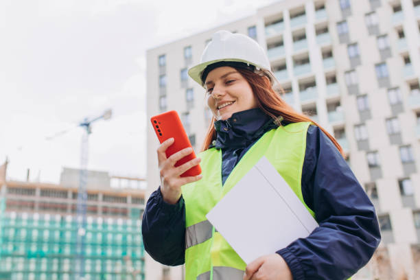 istock Happy Architect with a blueprints using smartphone at a construction site. Portrait of redhead woman constructor wearing white helmet and safety vest outdoors, technology or builing concept 1400812941