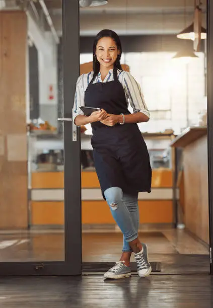 Portrait of businesswoman standing in her shop entrance. Young business owner using a digital tablet in her coffeeshop. Entrepreneur using an online app to order stock. Woman using wireless device