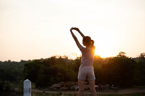 A woman standing with her back stretching after exercise against the sunset against the mountains in the background.