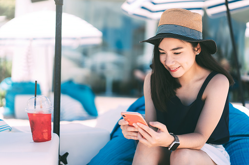 Summer travel holidays concept. Young adult traveller happy asian woman casual style outlook relax at outdoor blue color beach cafe on day. Using mobile phone for online application.