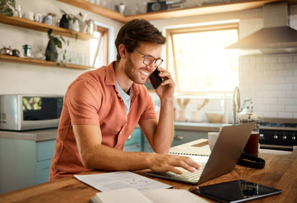 istock Young content caucasian businessman wearing glasses on a call using a phone while working on a laptop at home alone. Happy male businessperson smiling and talking on a cellphone while working in the kitchen at home 1400820516