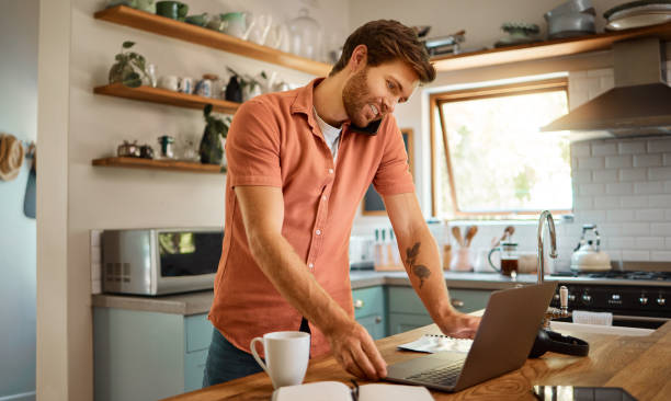 istock Young content caucasian businessman on a call using a cellphone while working on a laptop and drinking coffee at home alone. Happy male businessperson smiling and talking on a cellphone while working in the kitchen at home 1400820524