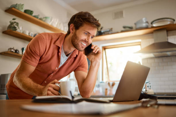 istock Young caucasian businessman on a call using a cellphone while working on a laptop and drinking coffee at home alone. Happy male businessperson smiling and talking on a cellphone while working in the kitchen at home 1400820526
