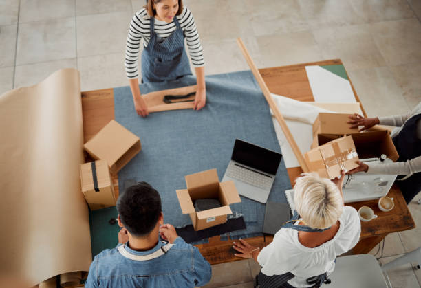 istock Four diverse clothing designers packing their products working together at a table at work. Tailors making packages while working together from above. Colleagues packing products into boxes at a shop 1400820580