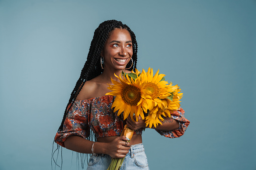 Happy african american young woman standing over blue background wearing summer look holding sunflower bouquet