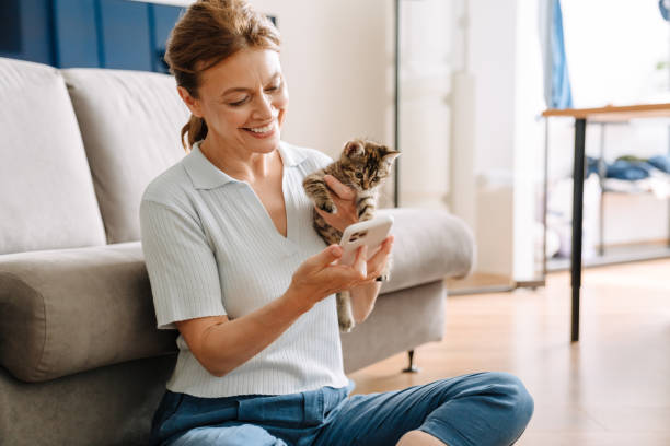 Senior woman holding kitten and using cellphone while sitting on floor