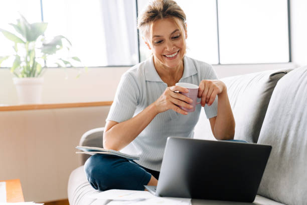 Ginger mature woman drinking coffee while working with laptop at home