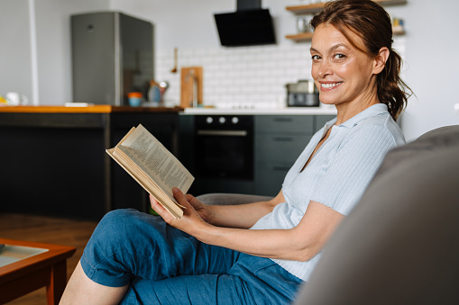 Ginger mature woman reading book while sitting on couch at home