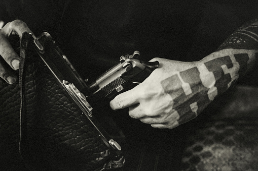 A man thoughtfully pulls a pistol out of an old lady's handbag with his tattooed one. He does this from a concealed position. Probably under the table.