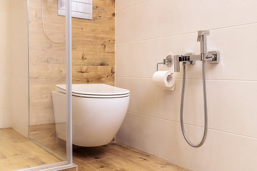 a white toilet and a hygienic shower for a bidet in the bathroom with ceramic tiles in natural colors like wood and white, in a Scandinavian style. Small bathroom interior. Close-up of plumbing.